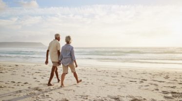 Rear View Of Senior Couple Walking Along Beach Hand In Hand